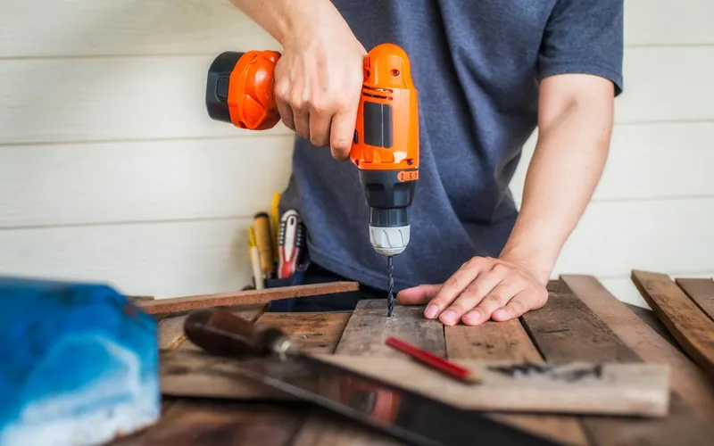Person drilling into a wooden board with a cordless drill at a workbench